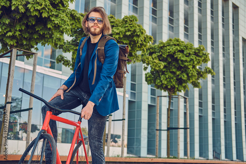 A handsome bearded male with long hair in stylish clothes and sunglasses with a backpack, riding a bicycle on the street, outdoors. © Fxquadro