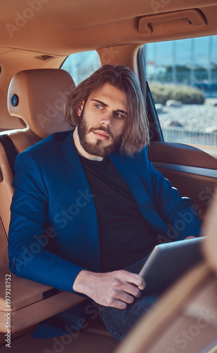 A handsome businessman with a beard and long hair sitting in the back seat of a luxury car and working with a tablet computer.