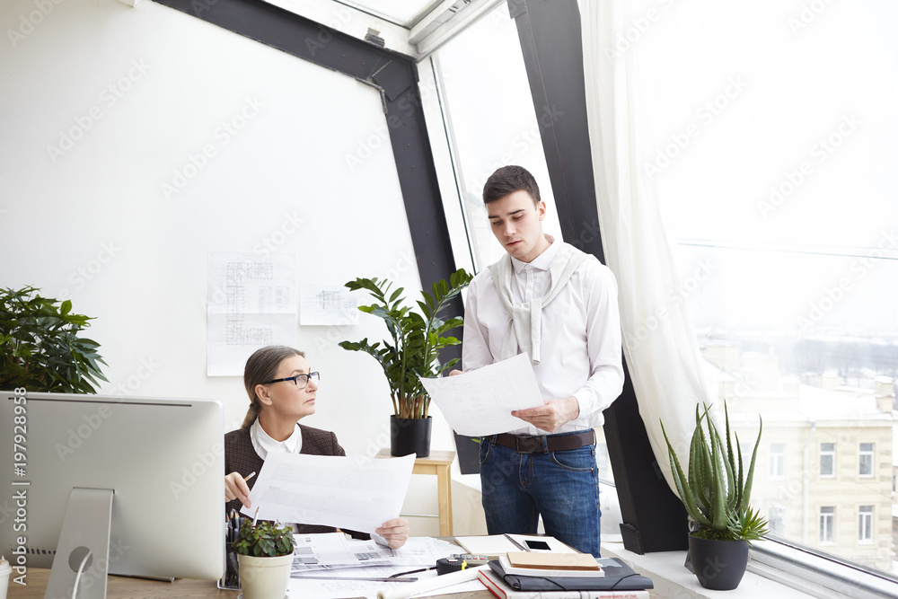 Portrait of two concentrated European constructors engineers senior woman and young man developing construction documentation, holding papers and having discussion in modern office interior Portrait of two concentrated European constructors engineers senior woman and young man developing construction documentation, holding papers and having discussion in modern office interior