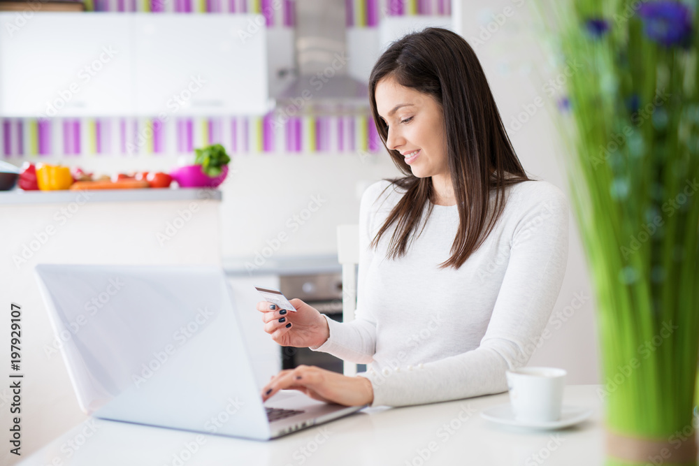 Young beautiful happy girl is typing her credit card info on a laptop while drinking coffee and sitting at the table of bright kitchen. Young beautiful happy girl is typing her credit card info on a laptop while drinking coffee and sitting at the table of bright kitchen.