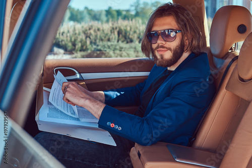 A handsome businessman with a beard and long hair sitting in the back seat of a luxury car and working with documents.