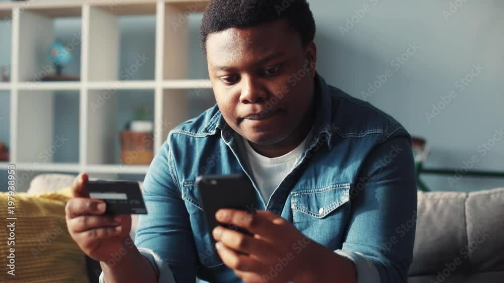 Concentrated serious young african american man use phone and credit card sit on sofa in home online shop work cell phone app authentic sunset guy portrait mobile smartphone slow motion rude close up Concentrated serious young african american man use phone and credit card sit on sofa in home online shop work cell phone app authentic sunset guy portrait mobile smartphone slow motion rude close up