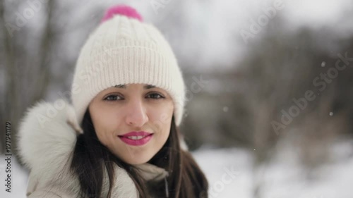 Closeup Brunette hair young women portrait with pink lips in beige down coat and hat smiling in snowy central park, snowfall.