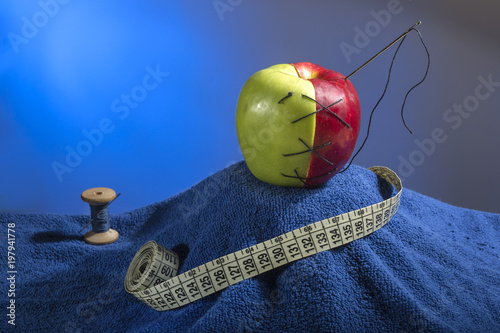 composition: green-red apple with the needle and thread on a blue towel. Near it blue coil of thread and centimeter for sewing photo