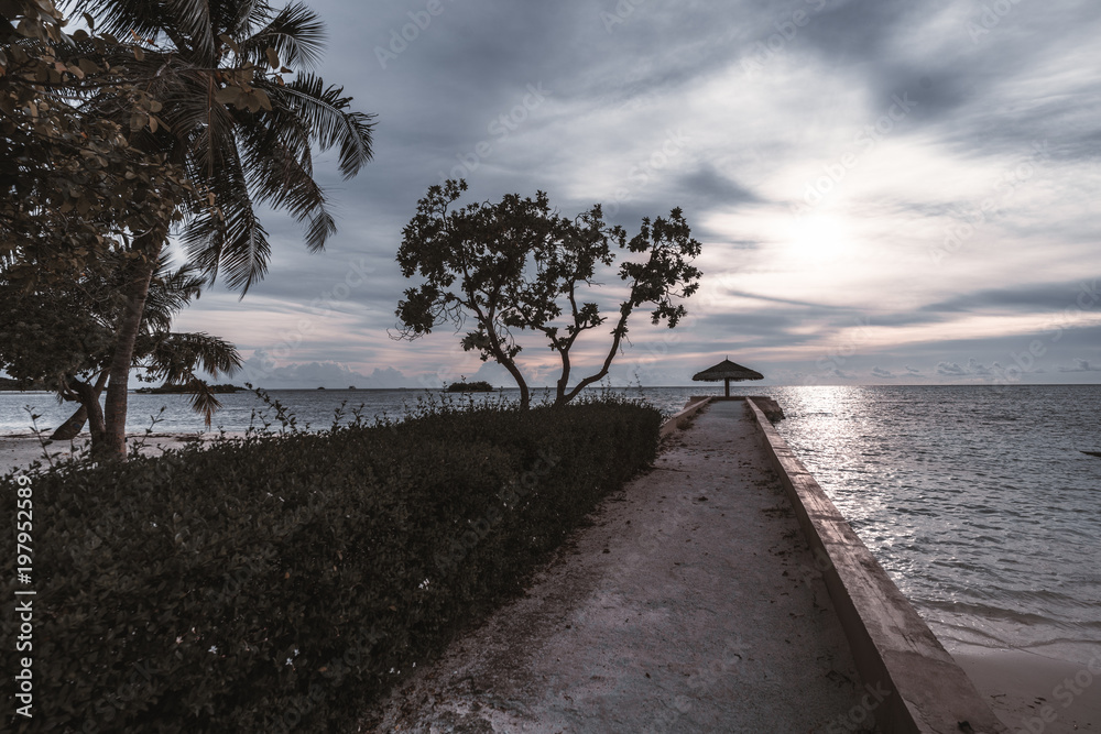 Long empty concrete pier with the sunshade at the end: palms, plants, and bushes on the left; beautiful lilac evening cloudscape, ocean and small islands in the background, Maldives