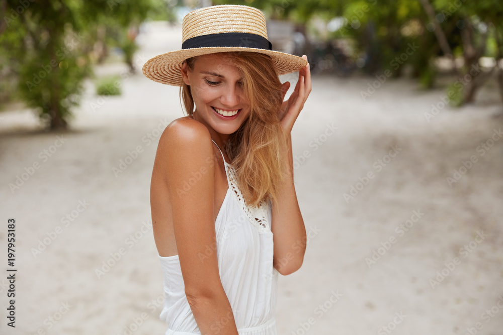 Positive shy female model in fashionable hat and dress, has pleasant smile, looks down with happy expression, poses outdoor against tropical background. Satisfied woman enjoys good summer rest