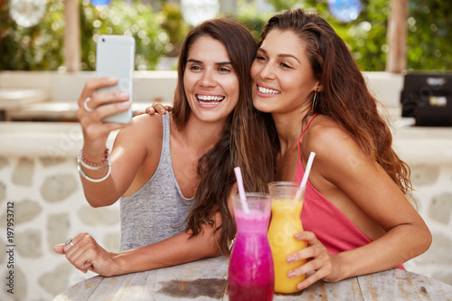 Beautiful female friends pose at camera of modern smart phone, make selfie, sit together in outdoor cafe, drink shake, have positive expressions. Pleased women colleagues meet after work for lunch