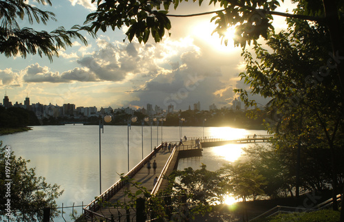 Represa municipal de São José do Rio Preto ao pôr do sol, com reflexos dourados na água, silhuetas de árvores, passarela com pessoas e céu parcialmente azul com nuvens photo