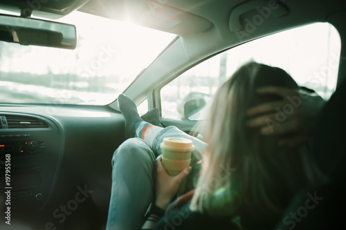 Woman is holding cup of coffee inside of car. Travel lifestyle. Legs on dashboard © Ivan Kurmyshov