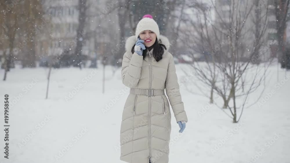 Brunette hair young women with pink lips in beige down coat and hat talking on the phone and smiling in snowy central park, snowfall. Slow motion. Brunette hair young women with pink lips in beige down coat and hat talking on the phone and smiling in snowy central park, snowfall. Slow motion.