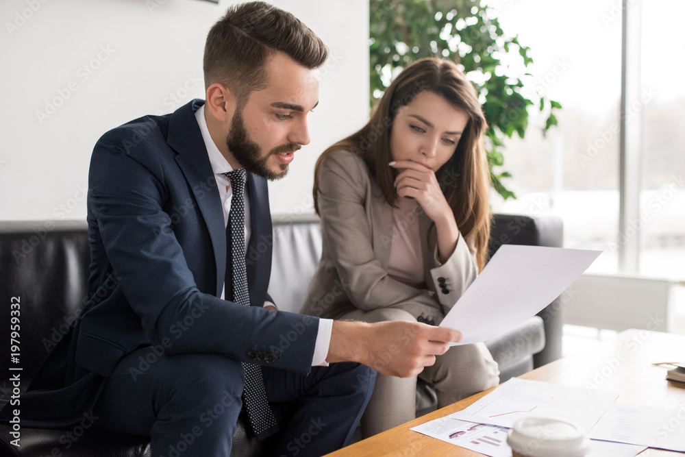 Portrait of two modern business people discussing documents in office, focus on pretty young woman reading contract Portrait of two modern business people discussing documents in office, focus on pretty young woman reading contract