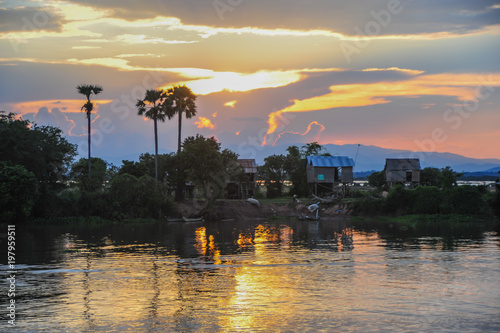 Abends am Mekong, Sonnenuntergang mit großer Farbpalette, Flussufer horizontal im Bild, Palmen am linken Uferrand, Hütten am rechten Rand, Farben der untergehenden Sonne spiegeln sich im Wasser photo