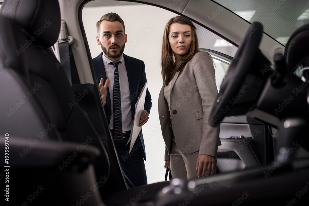 Portrait of handsome car salesman showing luxury car to woman in dealership showroom, shot from inside car Portrait of handsome car salesman showing luxury car to woman in dealership showroom, shot from inside car
