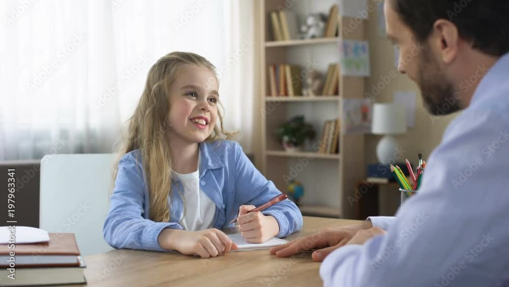 Smiling little daughter sitting at table and doing homework with father, care Smiling little daughter sitting at table and doing homework with father, care