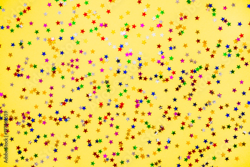 Scattered star shaped colorful glittering confetti over yellow background.