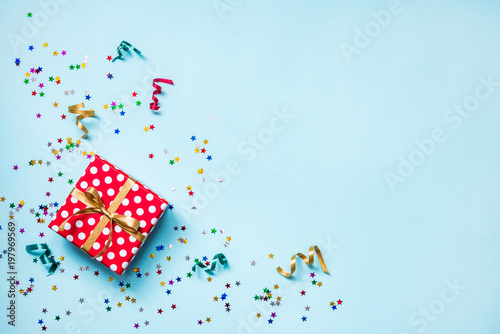 Top view of a red dotted gift box, scattered glittering star shaped confetti and colorful ribbons over blue background. Celebration concept. Copy space.