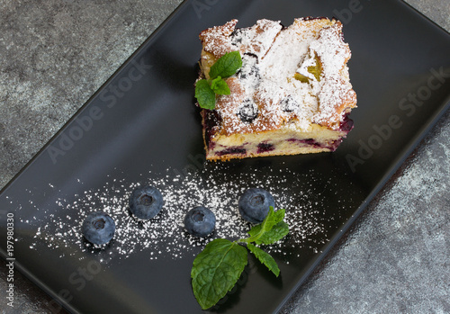 A piece of blueberry pie sprinkled with powdered sugar with blueberries and mint leaves on a black rectangular plate.