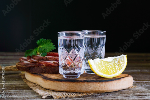 Two shot glasses of vodka with lemon slice and rye bread with salted bacon on the dark background. Traditional strong drink. Close-up.