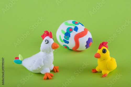 Close up of funny hen and chiken made of play clay sitting near big decorated egg on green background