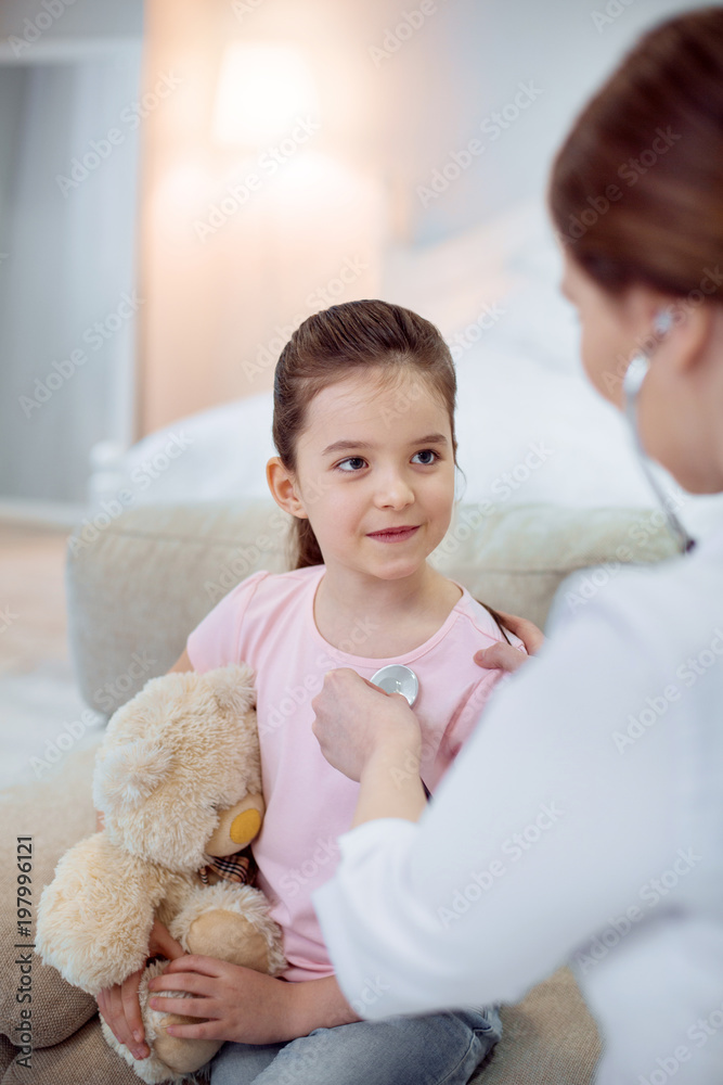 Childs needs. Appealing beautiful girl posing on blurred background and embracing plush bear while female doctor using stethoscope Childs needs. Appealing beautiful girl posing on blurred background and embracing plush bear while female doctor using stethoscope