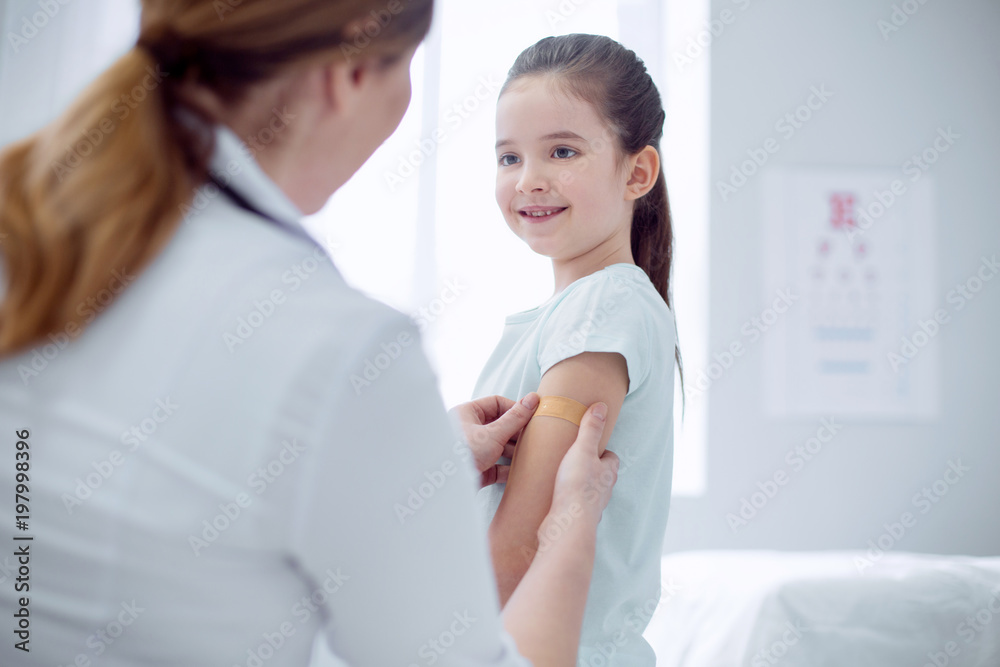 Doctors treatment. Jolly merry girl standing and smiling while doctor pressing patch against her arm Doctors treatment. Jolly merry girl standing and smiling while doctor pressing patch against her arm