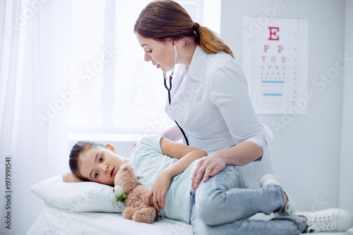 Everything ok. Worried skillful female doctor leaning over girl who lying and touching plush bear