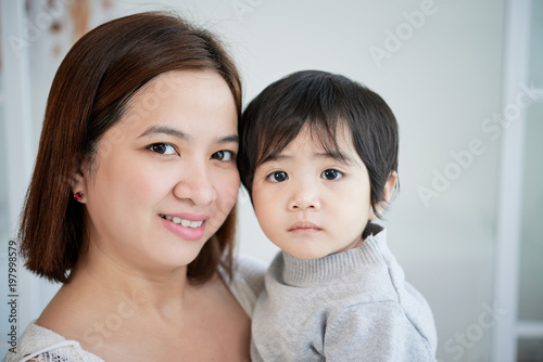 Head and shoulders portrait of beautiful Asian woman looking at camera with wide smile while holding her cute little son on hands  blurred background