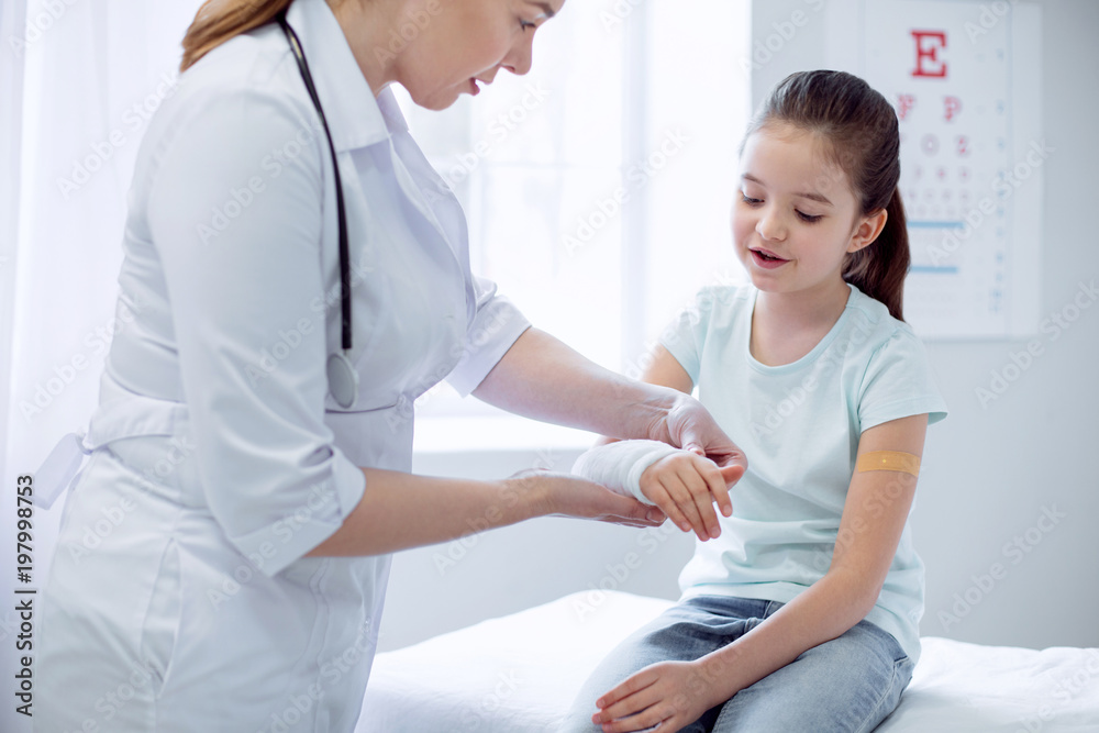 Broken arm. Charming smart interested girl broking arm and female doctor putting plaster while standing Broken arm. Charming smart interested girl broking arm and female doctor putting plaster while standing