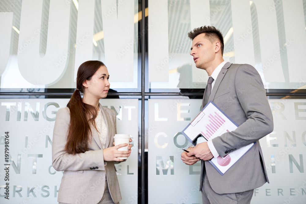 Waist up portrait of two business people, man and woman, talking to each other at coffee break standing in hall of modern office building Waist up portrait of two business people, man and woman, talking to each other at coffee break standing in hall of modern office building