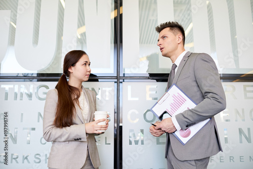 Waist up portrait of two business people, man and woman, talking to each other at coffee break standing in hall of modern office building