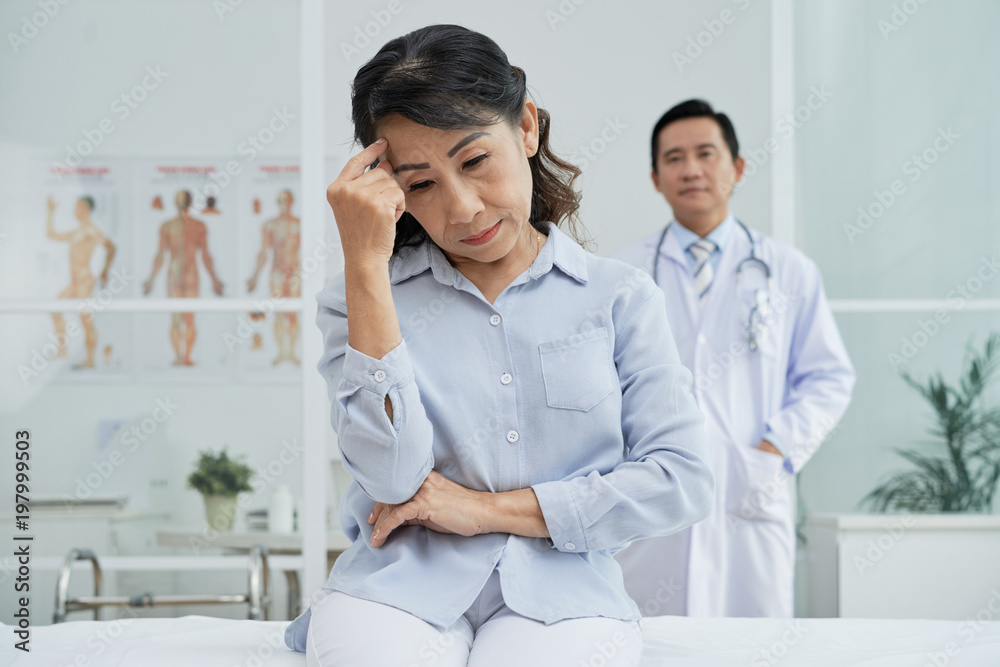 Sad Asian patient sitting on bed of modern ward and looking down while highly professional doctor wearing white coat standing behind her Sad Asian patient sitting on bed of modern ward and looking down while highly professional doctor wearing white coat standing behind her