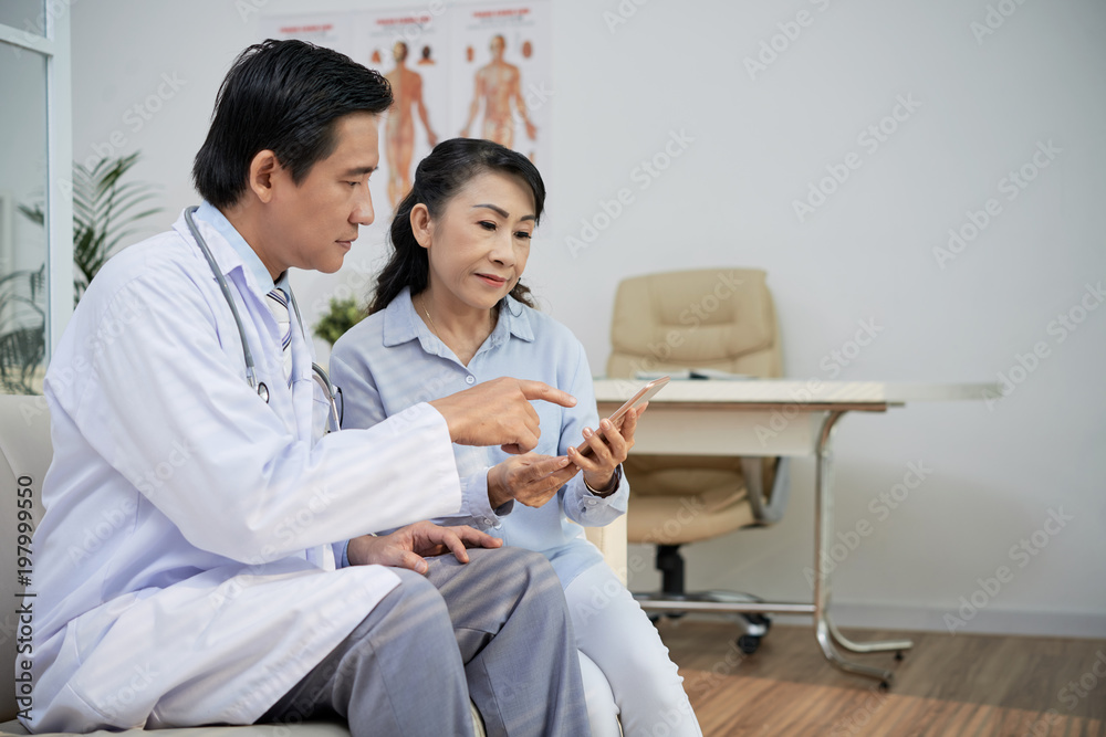 Obraz premium Confident Asian physician wearing white coat sitting on sofa and explaining senior patient how to use health monitoring application on smartphone, interior of modern office on background