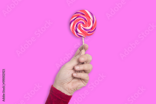 Left female hand holds colorful lollipop upright isolated on pastel pink background - concept lollipop sweets sugar health tast food kids family fun