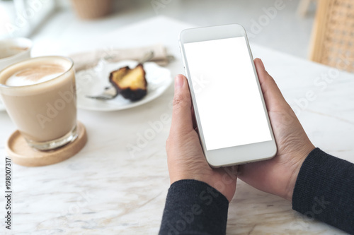 Mockup image of hands holding white mobile phone with blank screen with a cup of coffee and snack on table in cafe