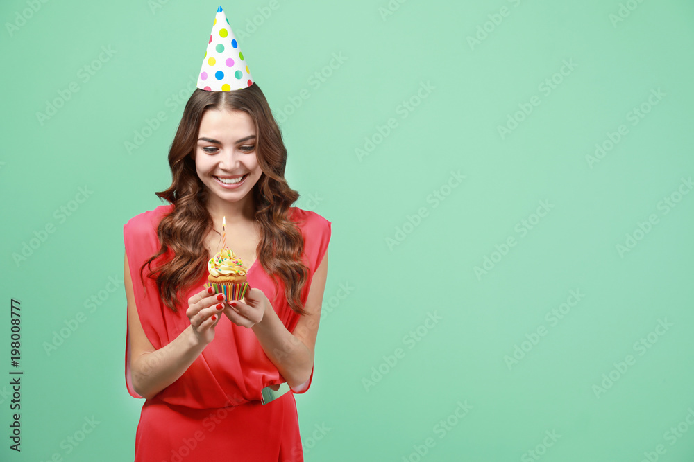 Happy young woman in party hat holding birthday cupcake with candle on color background Happy young woman in party hat holding birthday cupcake with candle on color background