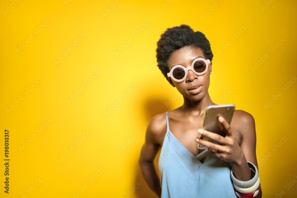 Fashionable girl standing in front a yellow wall, texting with her smart phone Fashionable girl standing in front a yellow wall, texting with her smart phone