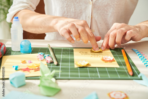 Close-up shot of young woman decorating handmade greeting card for her mother with paper flowers while sitting at wooden table of cozy living room photo