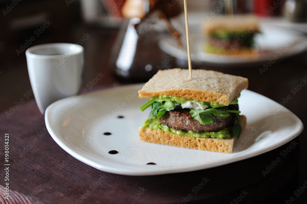 Burger with beef and greens in slices of unleavened bread. On white plate with drops of sauce. Coffee on background