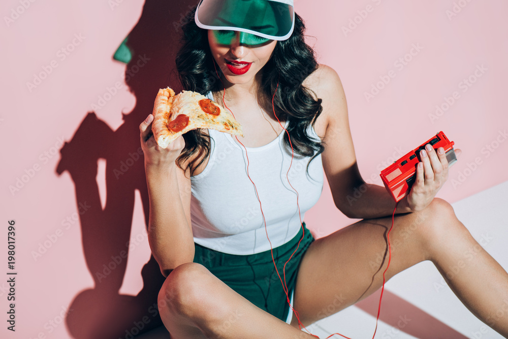 Φωτογραφία stylish woman in cap with retro music player holding piece of pizza on pink back