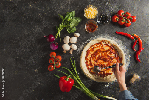 cropped shot of woman putting sauce on raw dough while cooking italian pizza on dark tabletop