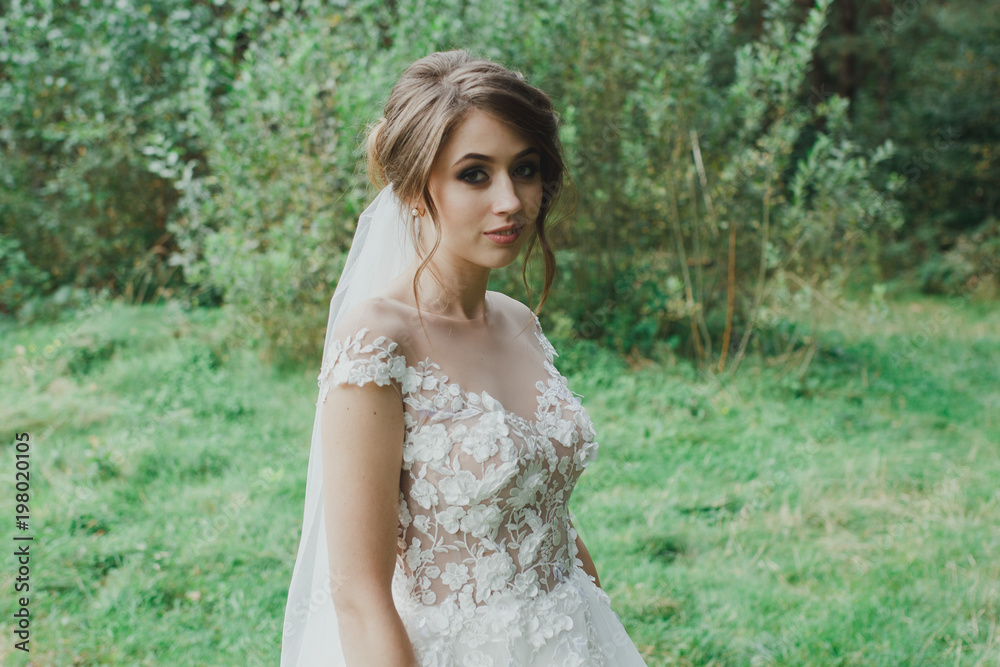 Beautiful bride with elegant simple hairdo is standing in the forest. Low open back white dress with flowers and tulle veil. Stylish hairstyle in outdoors romantic wedding photo. Bridal portrait.