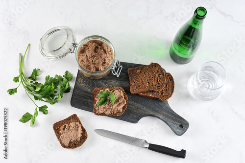 On a wooden board slices of black bread with liver pate. Next to the pot with pâté. In the frame, a knife, parsley, a bottle of water, a glass. Light surface. Macro photography. View from above.