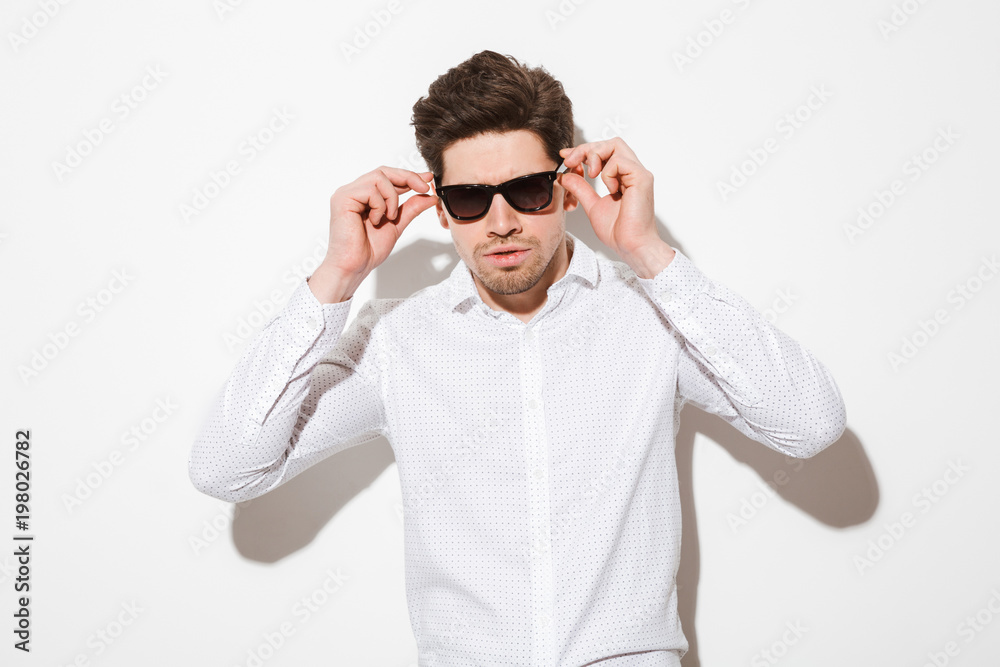 Fashion portrait of fancy man model dressed in touching sunglasses and looking on camera with serious gaze, over white with shadow Stock-foto | Adobe Stock
