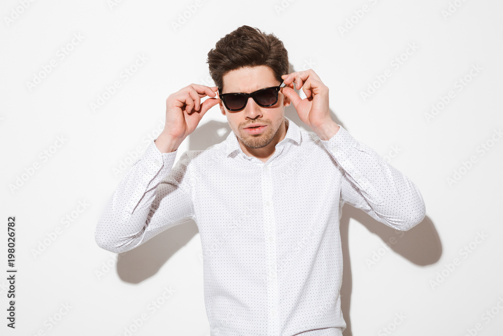 Fashion portrait of fancy man model dressed in shirt touching sunglasses and looking on camera with serious gaze, over white background with shadow Fashion portrait of fancy man model dressed in shirt touching sunglasses and looking on camera with serious gaze, over white background with shadow