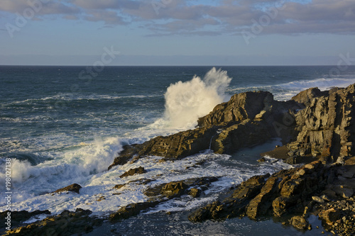 Felsige Meeresküste mit einer Welle die sich an einem Felsen hoch aufspritzend bricht.