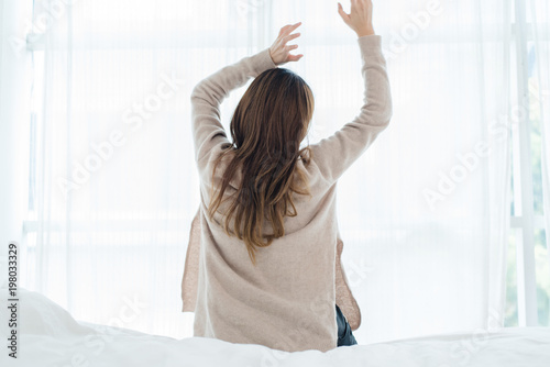 Back view of happy beautiful young Asian woman waking up in morning, sitting on bed, stretching in cozy bedroom, looking through window. Funny woman after wake up. She is stretching and smiling.