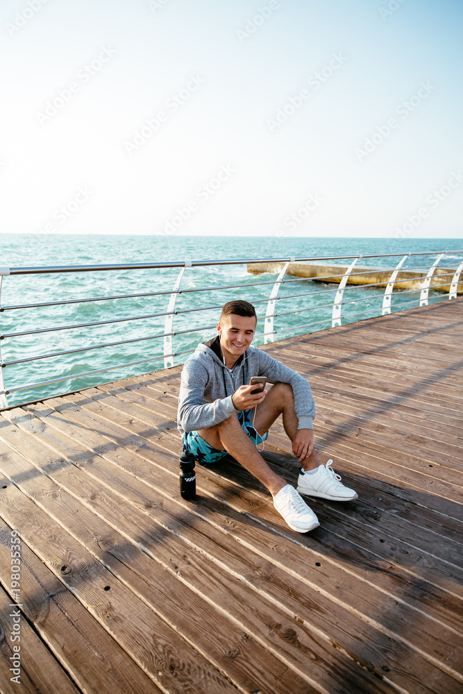 Cheerful guy in earphones using a smartphone, sitting on pier, by the sea