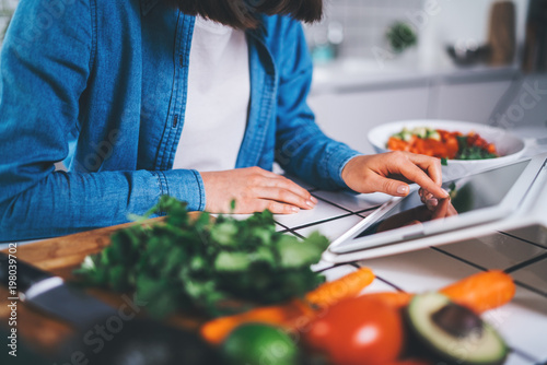 Closeup of female cook using digital tablet device for searching healthy meal recipes, young woman sitting in the kitchen preparing vegetarian food or salad, eating raw food
