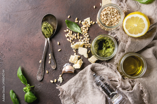 Traditional Basil pesto sauce in glass jar with ingredients above fresh basil, olive oil, parmesan cheese, garlic, pine nuts, lemon on linen cloth over dark texture background. Top view, space