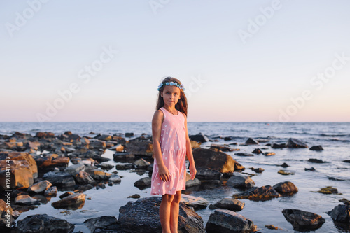 portrait of a cute girl in a pink dress and blue wreath standing near the water on sea shore.
